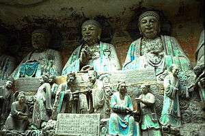 Stone sculptures of people interacting with each other and in the background a row of much larger seated buddha sculptures.