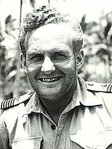 Informal head-and-shoulders portrait of grinning man in light-coloured shirt with palm trees in background