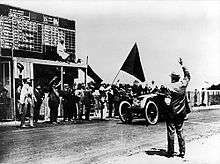 An open-topped sports car, with "3A" written on the front, crosses a white line painted on the track in front of a scoreboard, as a large black flag is waved and a crowd cheers.