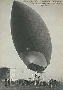 An airship is seen from below; mooring ropes are visible around the gondola; an airship's pointed bow looms at top left over the camera position, a slim, elegant, streamlined airship envelope recedes towards bottom right