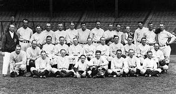 Three rows of men in white pinstriped baseball uniforms and one man in a dark jacket and white shirt stands at the left.