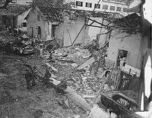  Soldiers with guns and civilians in white walk through the rubble of a white building demolished in an explosion. Pieces of wood, bent metal lie strewn on the ground. The roof has fallen off except for the support beams.