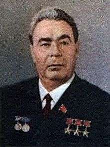 A portrait shot of an older man with greying hair, with five medals on his chest and one badge. He is wearing a black blazer over a white shirt and black tie.