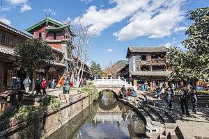 A small stone bridge across a small channel. On the opposite site there is a two storied red building with a three storied gate tower.