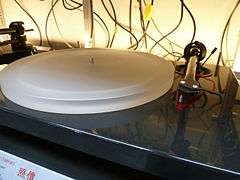 backlit image of a record player with grey plinth, a translucent acrylic platter, a black tonearm and a red phono cartridge