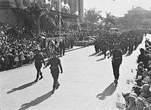 Soldiers in service dress march past a building in a city