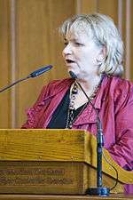 Head and upper body of a plump middle-aged woman with grey-blonde hair speaking at a wooden lectern, two slim microphones in front of her.