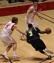 A basketball player in a dark blue uniform is dribbling past a defender in white.