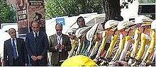 Six cyclists, wearing skin-tight jerseys, sit crouched over their bicycles in preparation to start a race. Men in suits are visible on the far side of the image.