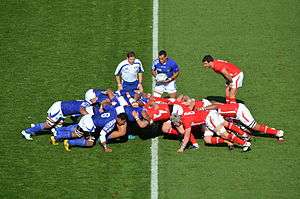 A player holds a ball in front of two opposing groups of eight players. Each group is crouched and working together to push against the other team.