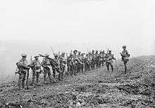 A line of soldiers in battle equipment face another soldier who is addressing them on a gentle slope. Behind them smoke or fog obscures the rest of the terrain.