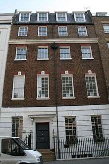 A terrace house with four floors and an attic. It is red brick, with a slate roof, and the ground floor rendered in imitation of stone and painted white. Each upper floor has four sash windows, divided into small panes. The door, with a canopy over it, occupies the place of the second window from the left on the ground floor.