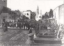 Towers and buildings some with people looking down from roofs with local people and soldiers in a large square with motor car in foreground