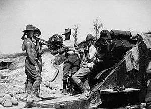 Bare chested men in slouch hats and breeches stand by a large artillery piece, ready to load a shell.