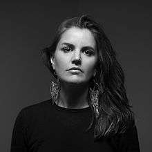 A 33-year-old woman is shown in upper body shot, taken in black and white. She has long dark hair swept over her right shoulder; and looks directly at the camera, wearing a simple black t-shirt and an elaborate metal earings. Background is dark grey.