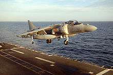 Grey jet aircraft executing a vertical takeoff from aircraft carrier at sea. Under each of the angled-down wings is an external fuel tank.