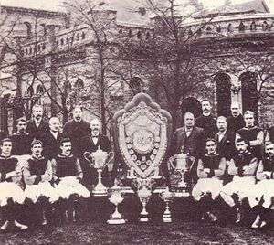 A sepia photograph with a large old structure in the background obscured by trees. In the foreground there is a large shield surrounded by five trophies. On either side of the shield stand 8 people.