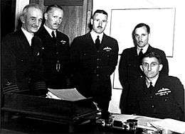 Five men in dark military uniforms behind a desk, four of whom are standing and one sitting