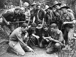 A group of soldiers kneel while another points to a mud model on the ground