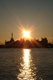 Sunset behind cluster of buildings, with water in foreground