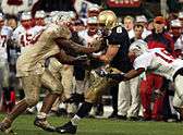 An American football player in a mud-covered blue and gold uniform is being tackled by two players in red and white uniforms as he catches the ball.