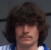 A headshot of a Caucasian man with long, shaggy brown hair. He also dons a mustache.