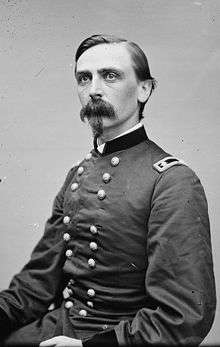 Head and shoulders of a thin white man with a bushy Van Dyke mustache and hair parted at the side. He is wearing a double-breasted military jacket with two stars on a patch on the shoulder.