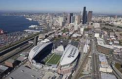 The stadium from the air on a clear day. SEAHAWKS STADIUM is painted on the white partial roof. The stadium is surrounded by roads and buildings.