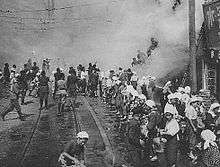 Black and white photo of women standing on a street passing buckets along a chain of people towards a building on fire. Other people are climbing a ladder from the street into the building.