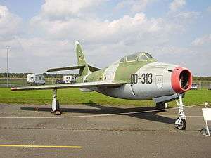 A camouflaged jet aircraft with a red ring around its nose intake, on display