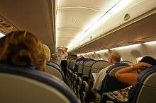 Interior of single-aisle jet aircraft, with people seated. Light illuminate the ceiling as a female stands near the front of aircraft