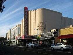 A color photograph of a movie theater facade and street scene. The theater is designed in the Streamline Moderne style, with Moorish elements, and a blade which reads "Alameda" standing vertically above a marquee announcing films.