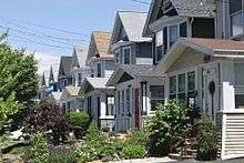 A row of detached wooden gabled houses on the right, all following a similar basic plan but of different colors, with small lawsn in front