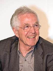 A smiling caucasian male with grey hair, blue eyes, in front of a white background. He is wearing a light grey shirt and dark grey jacket.