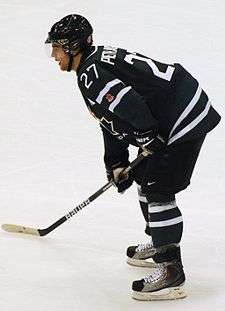 An ice hockey player leaning forward on his ice hockey stick and facing to the left of the camera. He is wearing a black helmet and uniform.