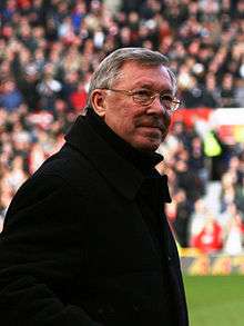 A white-haired man with glasses and a dark overcoat stands on the sideline of a stadium pitch. A stand full with people is visible in the background.