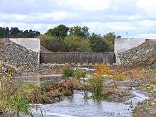 A muddy flow of water drops over an artificial concrete waterfall, from a tree-lined riverbed to a riprap-lined channel.