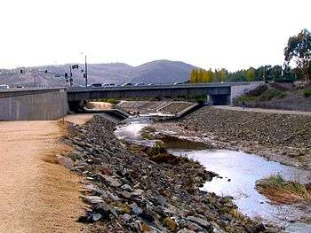 A channed stream between rocky banks runs towards a canyon in the distance as it flows under a concrete bridge