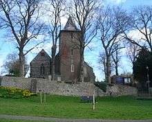 The tower and part of the body of a church viewed from a distance across a green, from which it is separated by a line of leafless trees and a low stone wall.