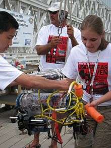 Two teenagers look down at a small vehicle. The vehicle is enclosed in a clear plastic tube and the machinery inside is clearly visible. To the right, a female student holds the vehicle. To the left, a male student adjusts tubing on the vehicle. In the back, a middle-aged man is holding a harness for the vehicle.