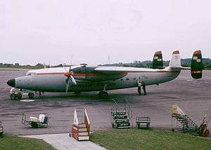 A twin-engine turboprop airliner with three fins parked on ramp while being serviced, with mobile staircases located nearby.