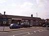 A long building with two chimneys protruding from its roof and four cars driving from left to right across a paved area in the foreground