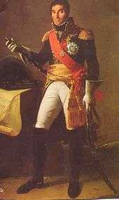 Formal full-length portrait of Masséna in dress military uniform, comprising white breeches with knee length black boots, dark cutaway coat with high collar and gold embroidery, a red shoulder sash and gold waist sash. He wears a large star of honour on his breast. He is a tall dark man with a long face and thick eyebrows. He looks quizzically at the observer and holds a marshal's baton, and sabre.