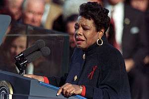 African American woman in her fifties, wearing a dark coat, standing at a podium reading to a crowd gathered behind her.