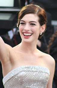 A broadly smiling young woman, wearing a white lame strapless gown and small earrings, waves presumably at a crowd. Her is parted on the left side and pulled up and back, off of her bare shoulders. The background is out of focus and unclear.