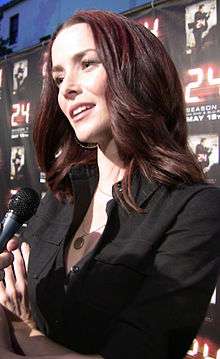 A 31-year-old woman with long, brown hair, smiling with her head tilted at someone holding a microphone to the left of the camera.