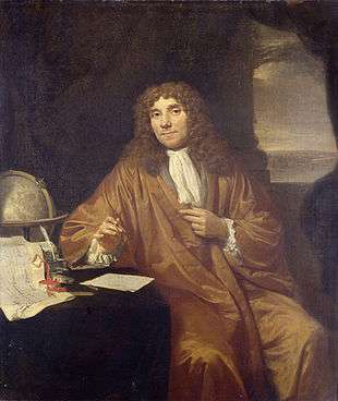 A man wearing a long, curly wig and a full robe is sitting, looking out. His left arm rests on a small table, with his left hand holding a box. Behind him is a globe.