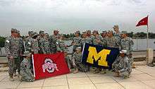 Two groups of soldiers in camouflage uniforms staring at each other on a waterfront; the left group carries a red flag with a silver "O" and the words "OHIO STATE" on it; the right group carries a blue flag with a yellow "M" on it.