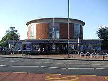 External view of a station building with a circular drum-shaped ticket hall with vertical panels of windows rising above a wider single storey entrance