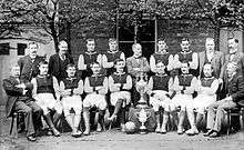 A group of men pose in two lines, one standing and the other seated. Eleven of the men are wearing dark-coloured football shirts with lighter sleeves, white shorts, shin pads and football boots. The remainder of the men are wearing formal suits. Almost all of the men sport moustaches. Displayed in the centre of the group are two large trophies.